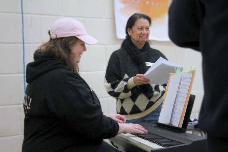 Two smiling women in a casual setting. One sits playing an electric keyboard, the other stands beside holding sheets of paper.