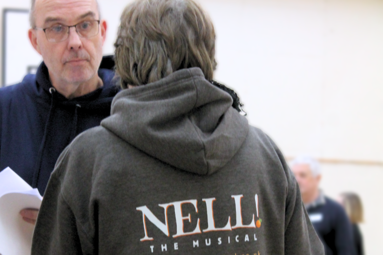 A man and a woman mid-conversation in a bright space. We see the back of the women who is wearing a Nell! The Musical hoodie.