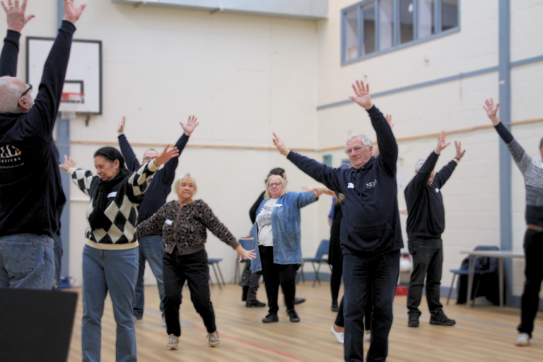 A group of people in a gymnasium setting are stretching their arms high.